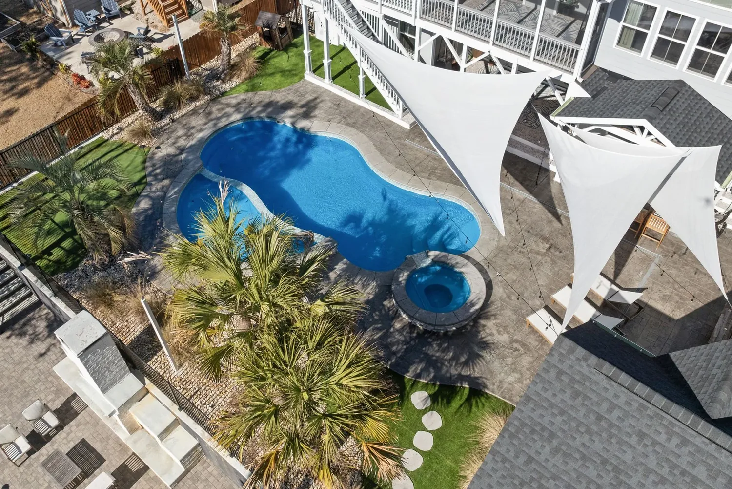 An aerial view of a freeform pool with a round spa, a stamped concrete deck, palms and shade sails