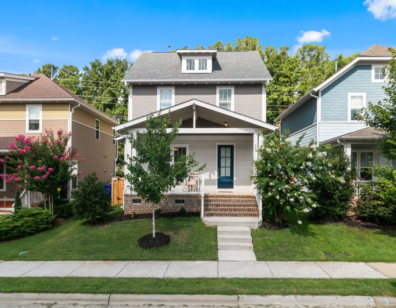 A two story house with a porch and a young tree in the front yard, between two neighbors