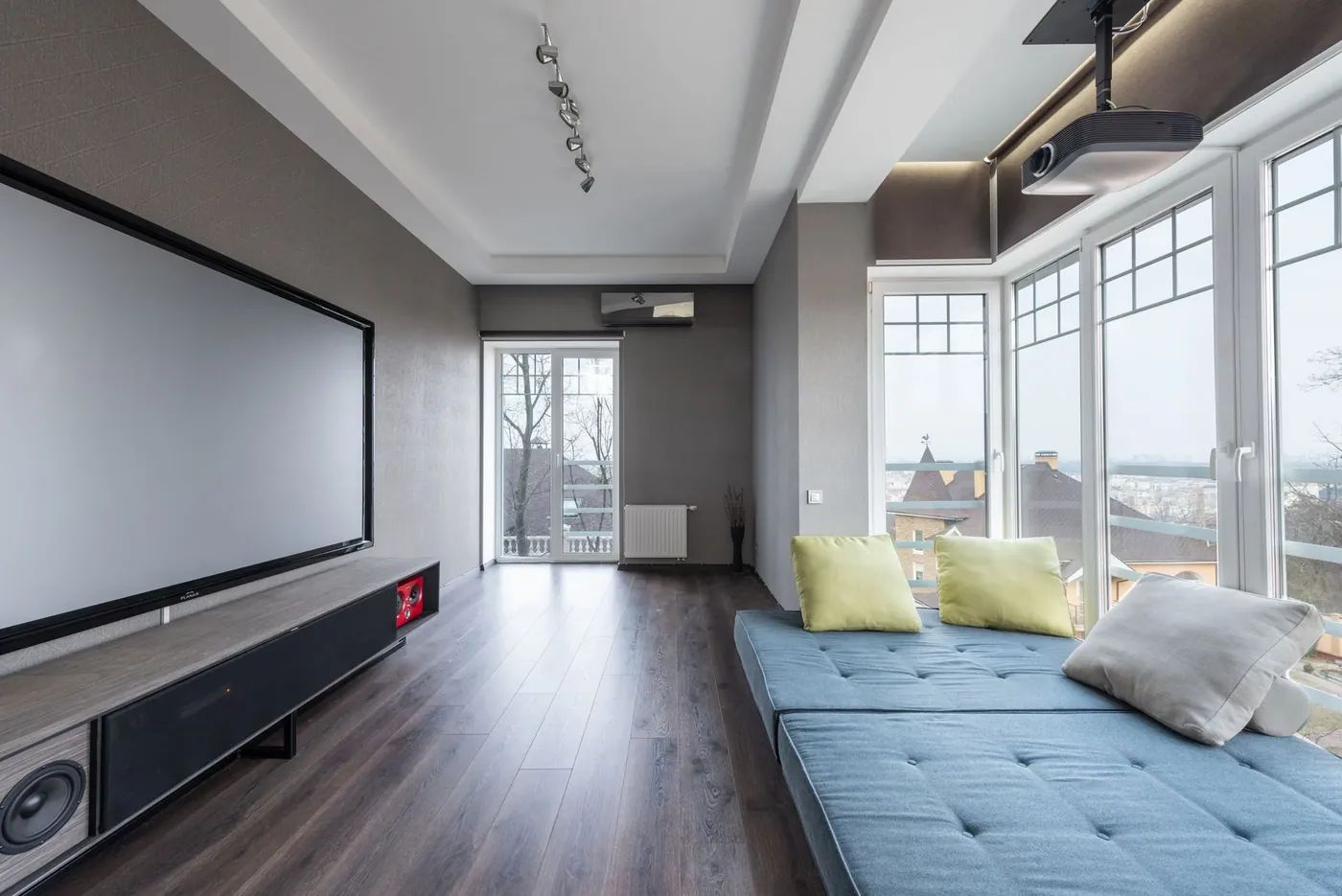 A bright media room with a large projector screen, a long low sofa and tall windows
