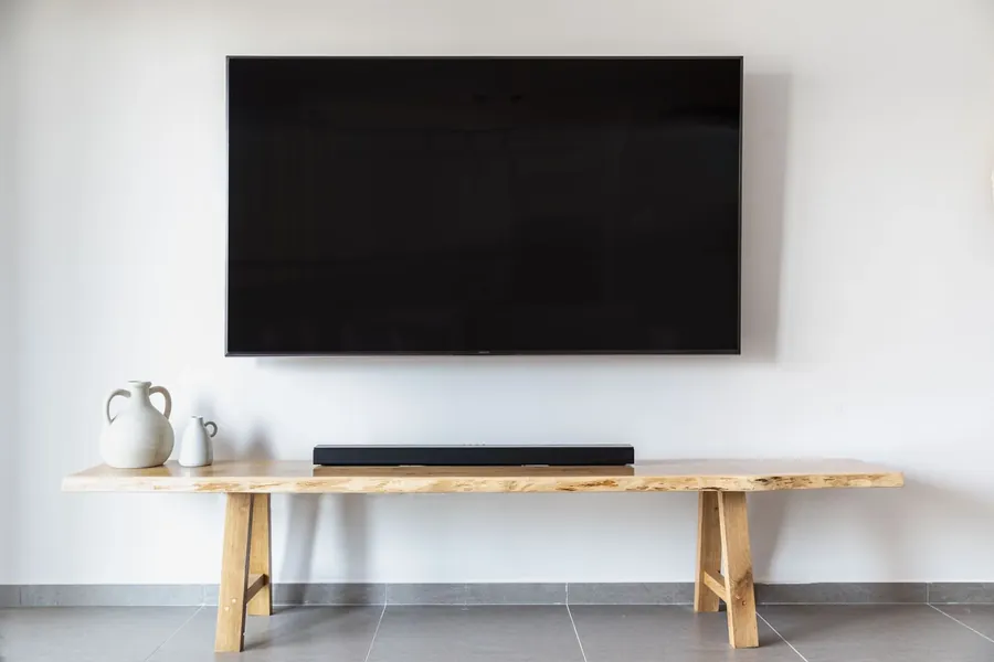 A wall mounted TV above a wooden bench with a soundbar, with no cables in sight