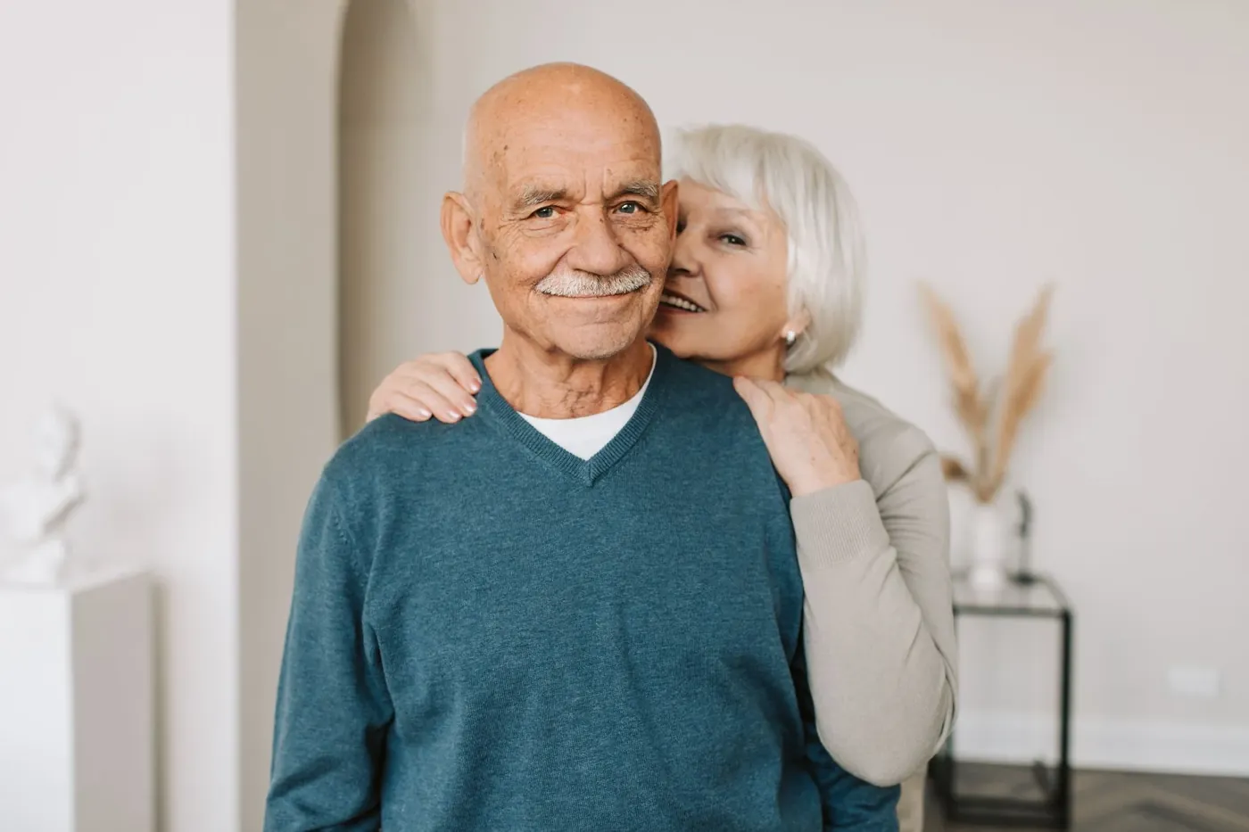An older man smiling at home with his wife's arms around his shoulders