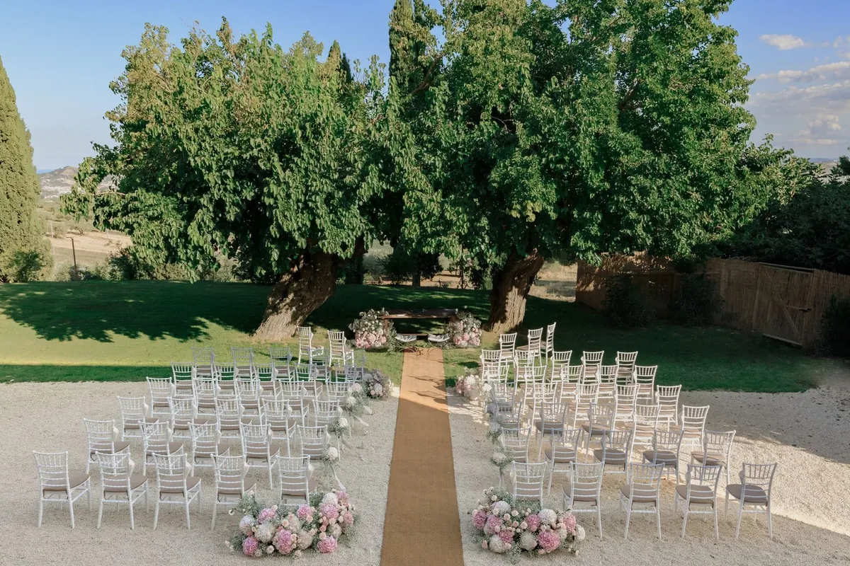 White chairs set out for a ceremony under big old trees, with flowers along the aisle
