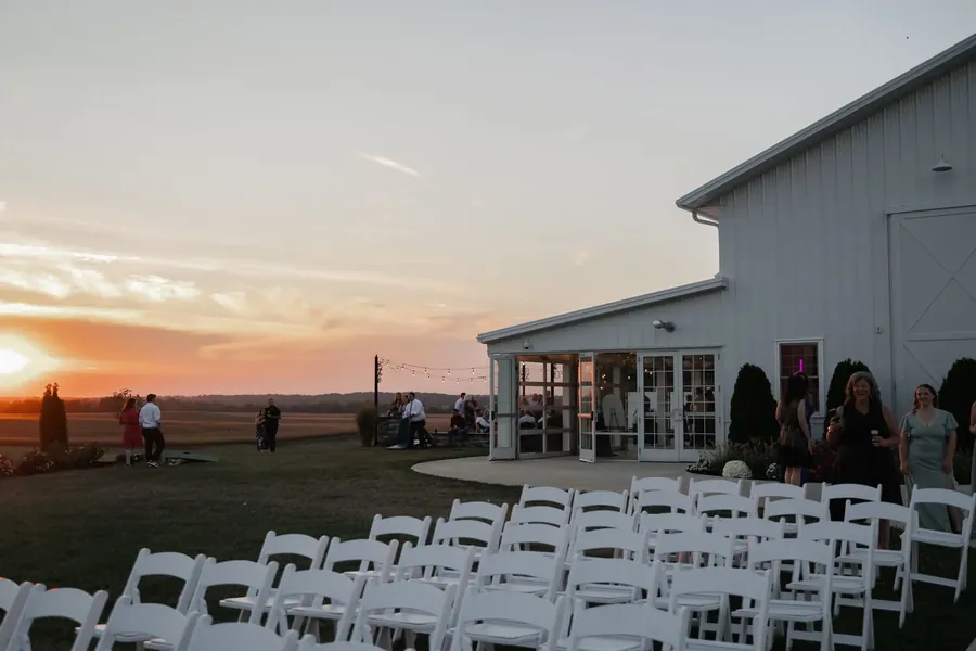 Guests on a terrace beside a white barn at sunset, with rows of white chairs