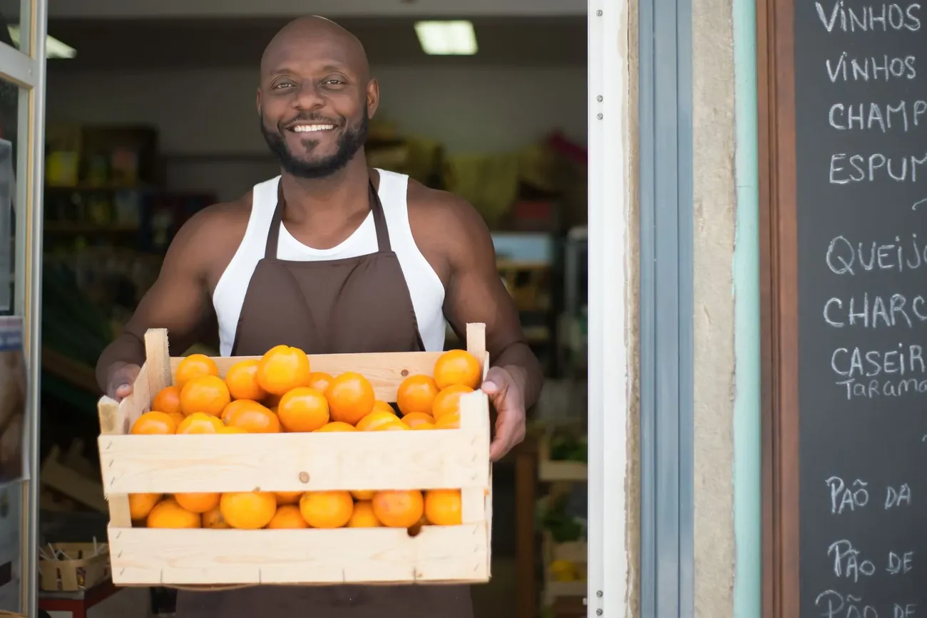 A smiling grocer in an apron holding a crate of oranges in his shop doorway