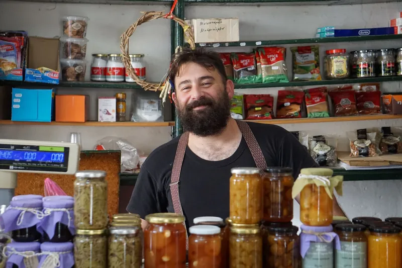 A bearded deli owner behind jars of preserves and a shop scale