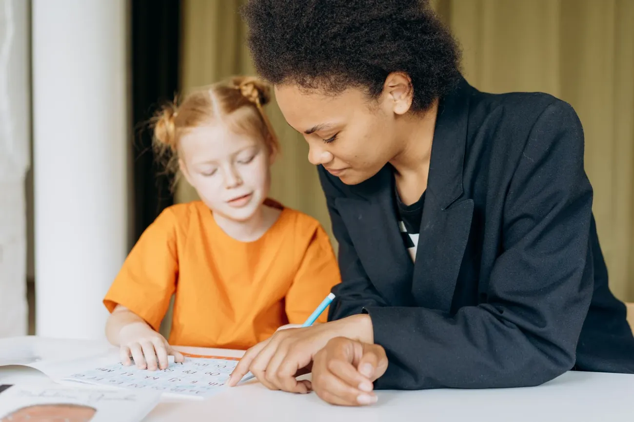 A tutor leaning in to help a young girl with red hair work through a worksheet