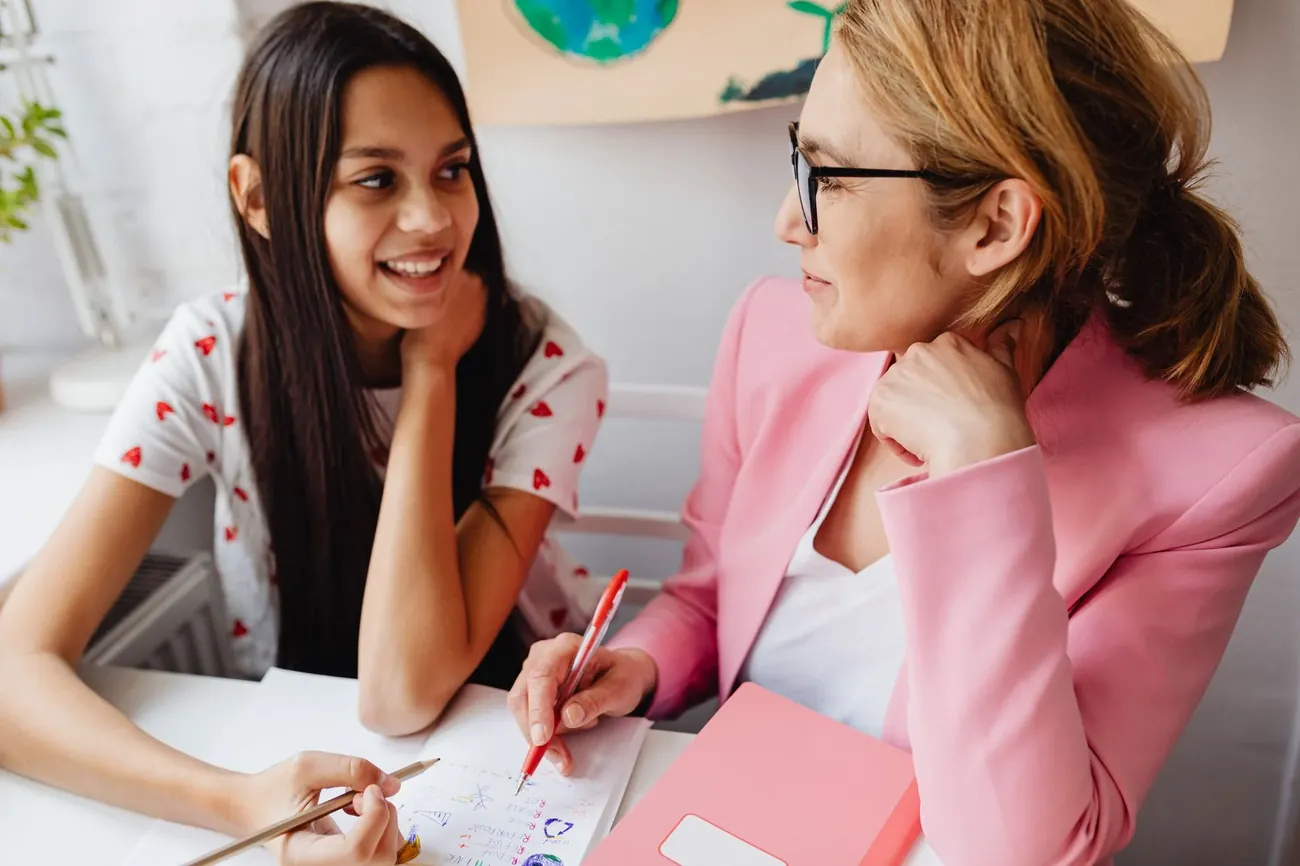 A smiling teenage girl working through a paper with her tutor, who holds a red pen
