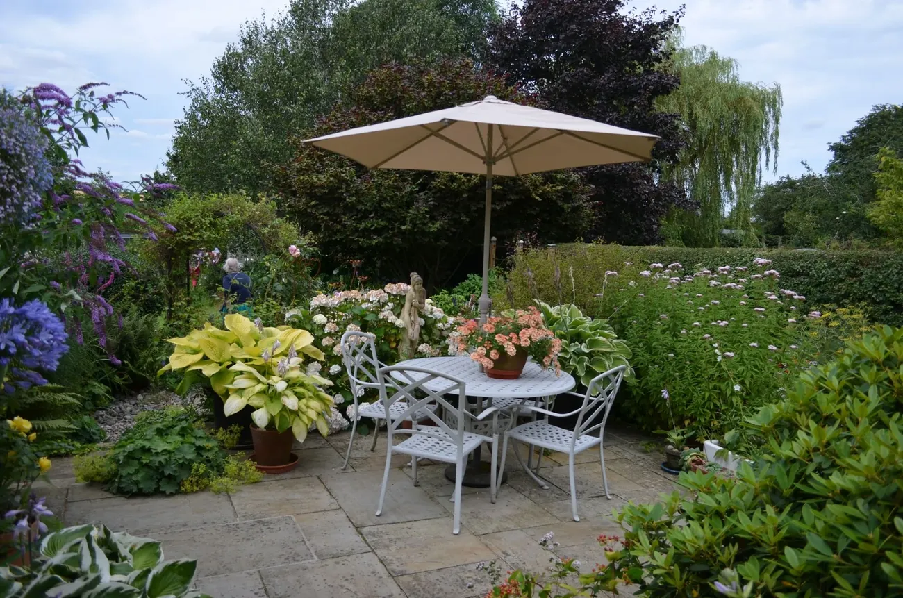 A stone patio with a parasol, white metal chairs and full borders