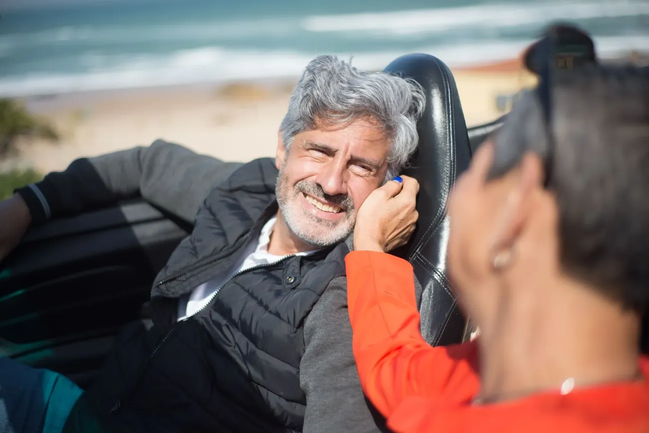 A smiling grey haired man leaning close as a woman speaks near his ear