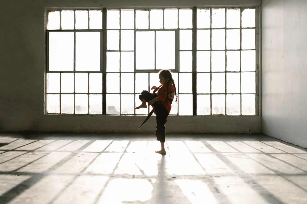 A student practising a kick alone in a studio lit by tall windows