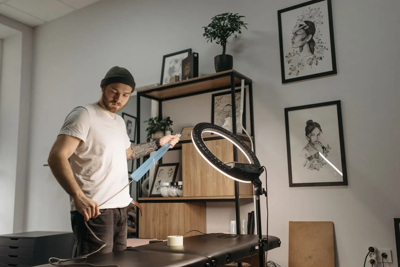 A tattoo artist standing in a bright studio with framed artwork and a ring light